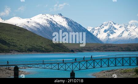 Personnes marchant sur le pont au-dessus du lac turquoise avec des montagnes enneigées en toile de fond. Tourné au lac Tekapo, Nouvelle-Zélande Banque D'Images