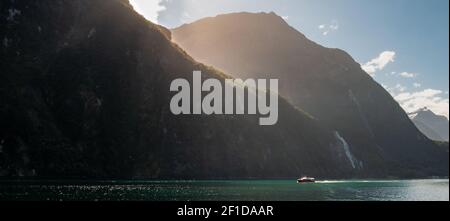 Photo panoramique du bateau naviguant dans le fjord par une journée ensoleillée avec des rayons du soleil créant une aura dorée autour du bord de la montagne. Milford Sound, Nouvelle-Zélande Banque D'Images