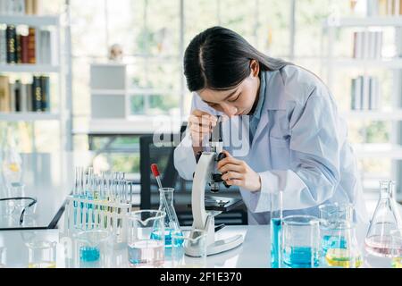 Jeune scientifique asiatique portant un blouse de travail médicale effectuant des recherches laboratoire tout en voyant les éléments à travers le microscope avec des équipements comme le test tube et flacon Banque D'Images