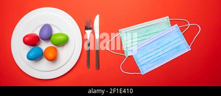 Fête de Pâques à la pandémie de coronavirus. Masques de protection Covid19 et table de Pâques sur fond rouge, vue de dessus. Banque D'Images