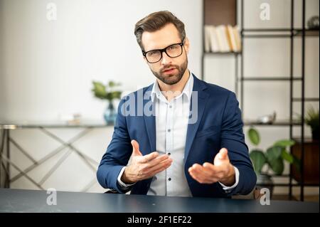 Jeune homme d'affaires plein de confiance en affaires vêtements décontractés regardant l'appareil photo, en présentant ses idées sur la nouvelle technologie ou le produit, en parlant et en faisant des gestes ou en invitant à rejoindre l'équipe Banque D'Images