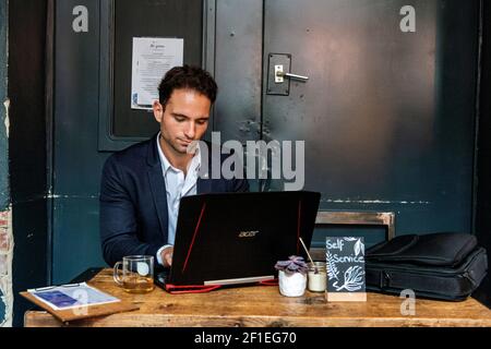 Berlin, Allemagne. Jeune adulte et professionnel gay travaillant avec son ordinateur portable et son smartphone à l'intérieur d'un café pour aider ses clients avec leur marketing en ligne et seo. Banque D'Images