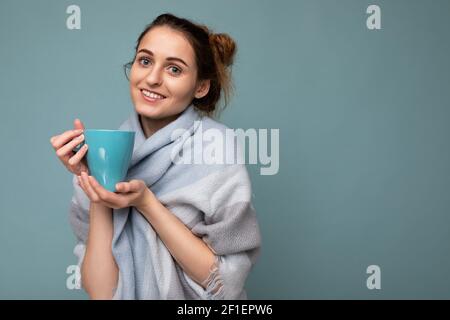 Photo d'une jeune femme brune souriante et belle portant chaude foulard bleu isolé sur fond bleu tenant une tasse bleue buvant café et Banque D'Images