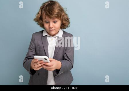 Photo d'un beau garçon surpris avec des cheveux bouclés portant du gris combinaison tenant et utilisant le téléphone isolé sur fond bleu regardant sur un smartphone Banque D'Images