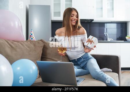 Belle femme dans un ensemble domestique boire du vin et ouvrir une boîte cadeau tout en célébrant l'anniversaire par appel vidéo. Jeune femme utilisant un ordinateur portable sans fil pour la fête en ligne à la maison. Banque D'Images