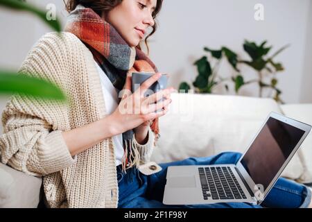 Jeune femme au repos regardant un ordinateur portable dans le confort de son appartement. Elle est assise sur un canapé, porte un foulard, boit un café. Vue latérale, rognée. Banque D'Images