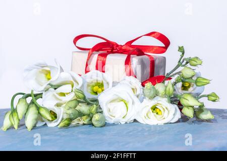 Petites roses blanches délicates et un cadeau blanc avec un ruban rouge sur fond bleu en bois, espace copie, vue de face. Carte postale parfaite pour la fête des mères, Banque D'Images