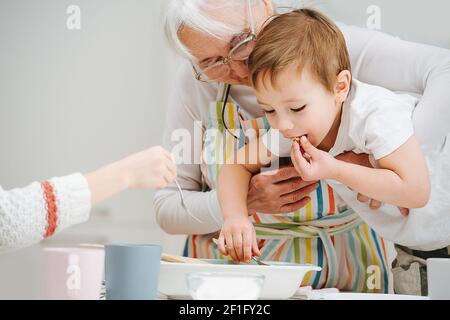 Frères et sœurs visitant leur grand-mère. Le garçon dans les bras de sa grand-mère a grimpé dans une assiette pour goûter la nourriture Banque D'Images