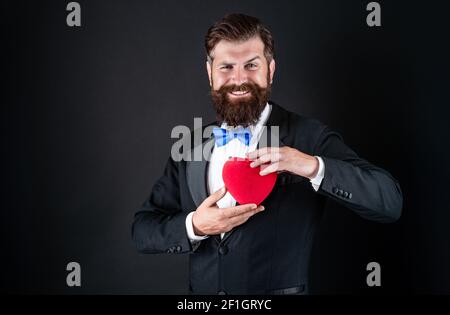 Dans l'amour. Beau et heureux homme de faire la proposition. Cadeau romantique. tuxedo homme avec coeur. Bonne Saint Valentin. Soyez ma Valentin. Amour et romance. Jeune homme barbu ayant l'air formel. 14 février. Banque D'Images