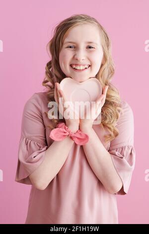 un enfant plein de sourire aux longs cheveux blonds ondulés avec une soucoupe en forme de coeur sur fond rose. Banque D'Images