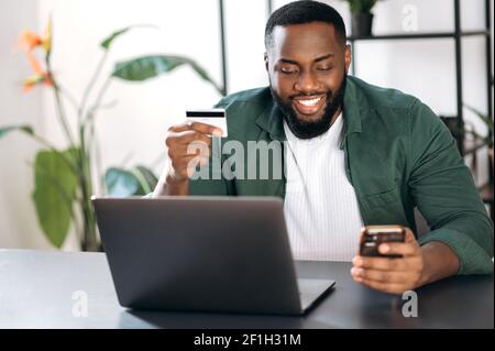 Souriant, un homme afro-américain élégant s'assoit à son bureau, à la maison ou au bureau, utilise son smartphone et sa carte de crédit pour le paiement en ligne ou les achats en ligne Banque D'Images