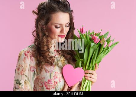 femme élégante et décontractée avec de longs cheveux de brunette ondulés avec bouquet de tulipes et coeur rose isolé sur fond rose. Banque D'Images