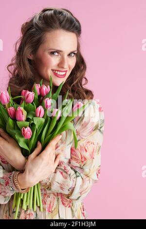 femme élégante et souriante d'âge moyen en robe fleurie avec bouquet de tulipes sur fond rose. Banque D'Images