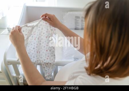 Vue arrière sur une femme de race blanche inconnue portant des vêtements pour bébé Par le berceau à la maison - femme enceinte préparant la combinaison-pantalon pour bébé dans une pièce lumineuse - mater Banque D'Images