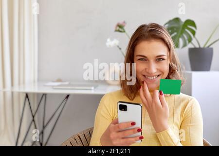 Elle est ravie de la femme avec une carte en plastique et un smartphone pour l'achat en ligne lorsque vous regardez l'appareil photo à la maison Banque D'Images