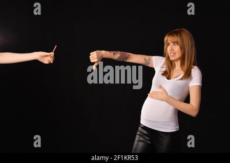 La jeune femme enceinte refuse de prendre la cigarette suggérée sur fond sombre. Concept de mode de vie sain Banque D'Images