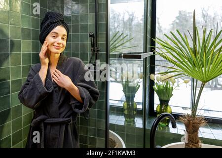 Femme en robe avec cheveux enveloppés dans une serviette après la douche dans la belle salle de bains verte intérieure. Sourire à l'appareil photo. Concept beauté et soin. Banque D'Images
