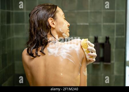Femme souriante prenant la douche et sochant le corps avec gel douche dans la salle de bains verte. Tenir le débarbouillette à la main. Prendre des photos quotidiennes. Vue arrière. Banque D'Images