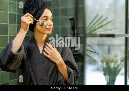 Femme massant son visage avec un rouleau de pierre du visage, en robe avec cheveux enveloppés dans une serviette après la douche dans la salle de bains verte à la maison. Regardez sur le côté. Concept beauté et soin. Banque D'Images