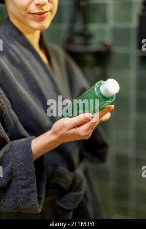 Femme après la douche tenant et regardant sur une bouteille avec lotion verte dans la salle de bains. Concept beauté et soin. Produits de soins pour les cheveux et le corps. Banque D'Images