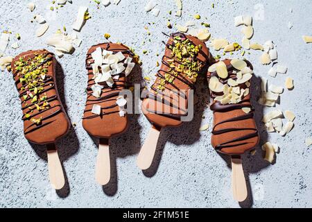 Glace au chocolat popsicle parsemée d'amandes, de noix de coco et de pistaches sur fond gris. Glace végétalienne au chocolat noir, vue du dessus. Banque D'Images