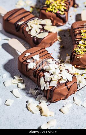 Glace au chocolat popsicle parsemée d'amandes, de noix de coco et de pistaches sur fond gris. Glace végétalienne au chocolat noir, vue du dessus. Banque D'Images