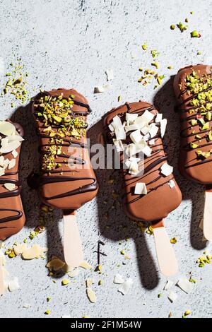 Glace au chocolat popsicle parsemée d'amandes, de noix de coco et de pistaches sur fond gris. Glace végétalienne au chocolat noir, vue du dessus. Banque D'Images