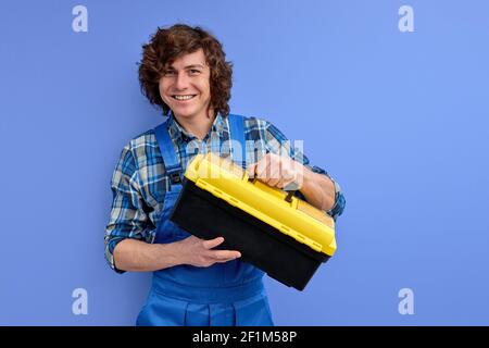 Jeune homme de construction en vêtements de travail tient la boîte à outils dans les mains et regarde la caméra souriante isolée sur fond violet. Constructeur portrait de constructeur Banque D'Images