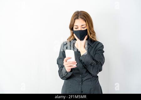 Jeune femme modèle isolée sur fond blanc vêtue d'affaires sombre le maillot et le masque de protection permettent à votre menton de rester inquiète et de regarder un smartphone lecture de lates Banque D'Images