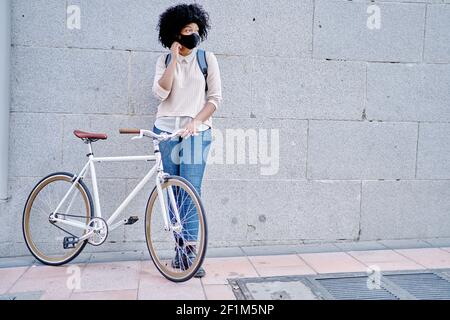 Femme afro-américaine avec un smartphone dans la main. Femme avec masque de protection. Vélo dans la ville. Photo de haute qualité Banque D'Images