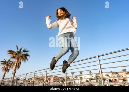 Bonne fille souriante sautant dehors haut avec les bras vers le haut regardant vers le bas l'appareil photo avec ses cheveux rouffés par le vent. Concept de bonheur et de bonne vie Banque D'Images