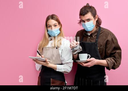 Les jeunes serveurs prêts à servir les clients dans un café ou un restaurant, se posent à la caméra isolée sur fond rose, le serveur mâle versant le café dans le blanc Banque D'Images