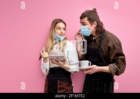 deux jeunes serveurs en tablier et masque discutent des commandes, prêts à servir les clients pendant la quarantaine, serveuse amicale écrivant des notes et parlant Banque D'Images