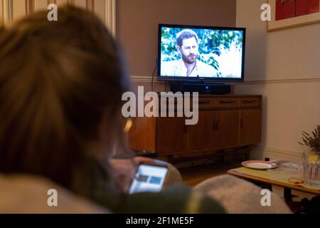 Vue de l'arrière, une jeune femme regarde l'écran sur son téléphone tout en regardant le duc et la duchesse de l'interview télévisée controversée de Sussex avec la reine Oprah, émission de tchat américaine, diffusée à ITV au Royaume-Uni, le 8 mars 2021, à Londres, en Angleterre. L'interview cntroversiale a accusé les membres de la famille royale britannique de racisme, obligeant le couple à cesser les fonctions officielles et à renoncer à leurs titres royaux. Banque D'Images