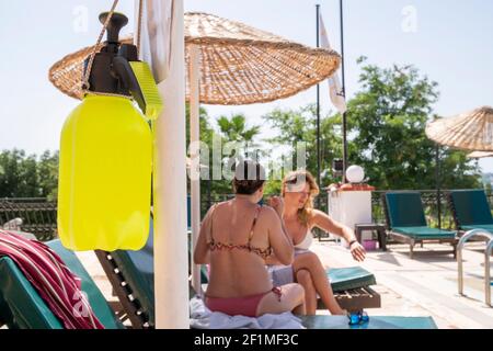 Été 2021 - Nouvelle norme. Désinfectant à base d'alcool contre Covid-19 suspendu à un poteau dans la zone de la piscine. Deux femmes assises sur des chaises longues et parlant Banque D'Images