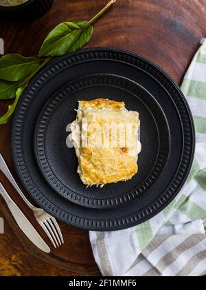 Délicieux lasagnes quatre fromages sur des assiettes en céramique noire, des feuilles vertes, des couverts et un torchon à base de bois. Gros plan, vue de dessus. Banque D'Images