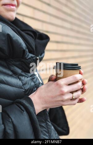 vue verticale de la tasse de café jetable par les mains d'une jeune femme caucasienne méconnaissable. Elle s'habille d'un élégant manteau noir et se tient près d'un mur de briques Banque D'Images