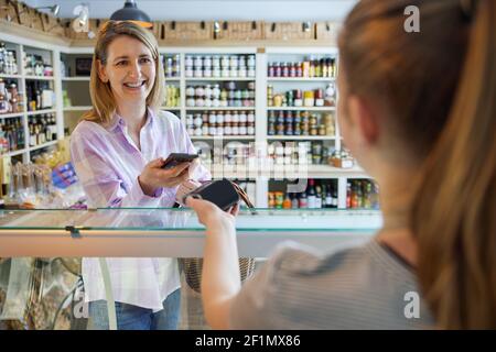 Client Femme souriant Delicatessen Food Store paiement sans contact avec Téléphone mobile pour le shopping au vendeur Banque D'Images