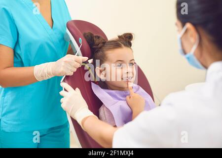 Une petite fille dans un cabinet dentaire montre deux femmes dentistes une dent qui l'inquiète. Banque D'Images