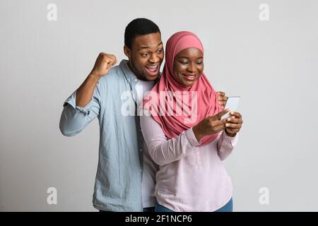 Couple noir musulman excité regardant l'écran de téléphone mobile, utilisant une nouvelle application fraîche, fond gris studio. Homme et femme afro-américains joyeux Banque D'Images