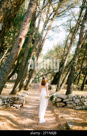 La mariée marche le long du chemin parmi les arbres dans la plantation et tient un bouquet, vue arrière Banque D'Images