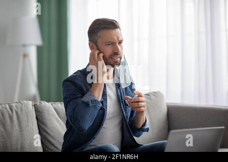 Homme d'âge moyen barbu qui retire son masque de protection, grattant sa barbe qui démange, travailleur indépendant travaillant sur un ordinateur portable à la maison pendant une pandémie de coronavirus, Banque D'Images