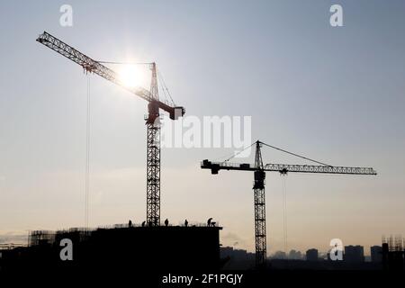 Silhouettes de grues de construction et de travailleurs sur le bâtiment résidentiel non fini contre le ciel et le soleil éclatant. Construction de logements Banque D'Images