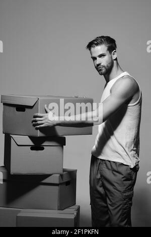 Guy se tient parmi les boîtes, l'espace de copie. Homme avec barbe est sur fond gris foncé. Concept de livraison et de déménagement. Macho au visage sérieux porte un t-shirt blanc. Banque D'Images