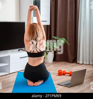 Femme sportive faisant du yoga de forme physique à la maison via un ordinateur portable par appel vidéo en ligne à distance. Jeune femme perdant du poids par l'entraînement en ligne à distance de gym. Vue arrière unr Banque D'Images