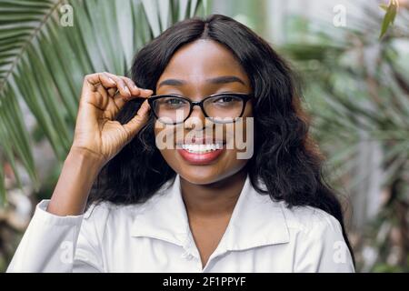 Jolie femme afro-américaine heureuse agronome, botaniste, portant des lunettes et un manteau blanc de laboratoire, travaillant en serre et regardant la caméra. Gros plan Banque D'Images