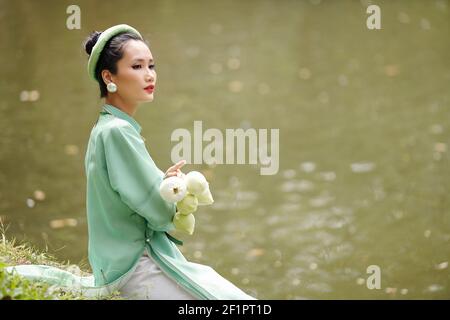 Jolie jeune femme vietnamienne pensive en robe traditionnelle et tête de lit assis au bord de la rivière avec des fleurs de lotus entre les mains Banque D'Images