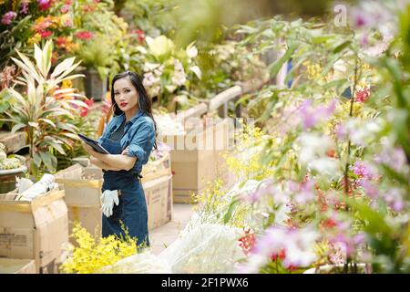 Portrait d'une jeune femme asiatique en tablier en denim debout dans un jardin de pépinière avec tablette numérique en mains Banque D'Images