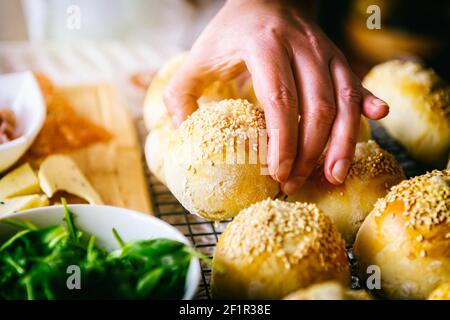 Petits pains faits maison aux graines de sésame. Une main de femme atteint pour un rouleau, une table avec le petit déjeuner Banque D'Images