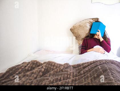 une jeune femme lit allongé dans un lit avec un livre bleu Banque D'Images
